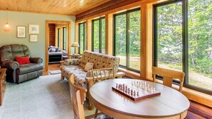 A cozy room with a wooden ceiling, a sofa, a chair, a table with a chessboard, and large windows offering a view of the outdoors.