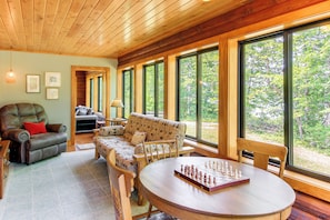 A cozy room with a wooden ceiling, a sofa, a chair, a table with a chessboard, and large windows offering a view of the outdoors. - Serene Wilderness Acres Retreat on Bear Lake (McGrath)