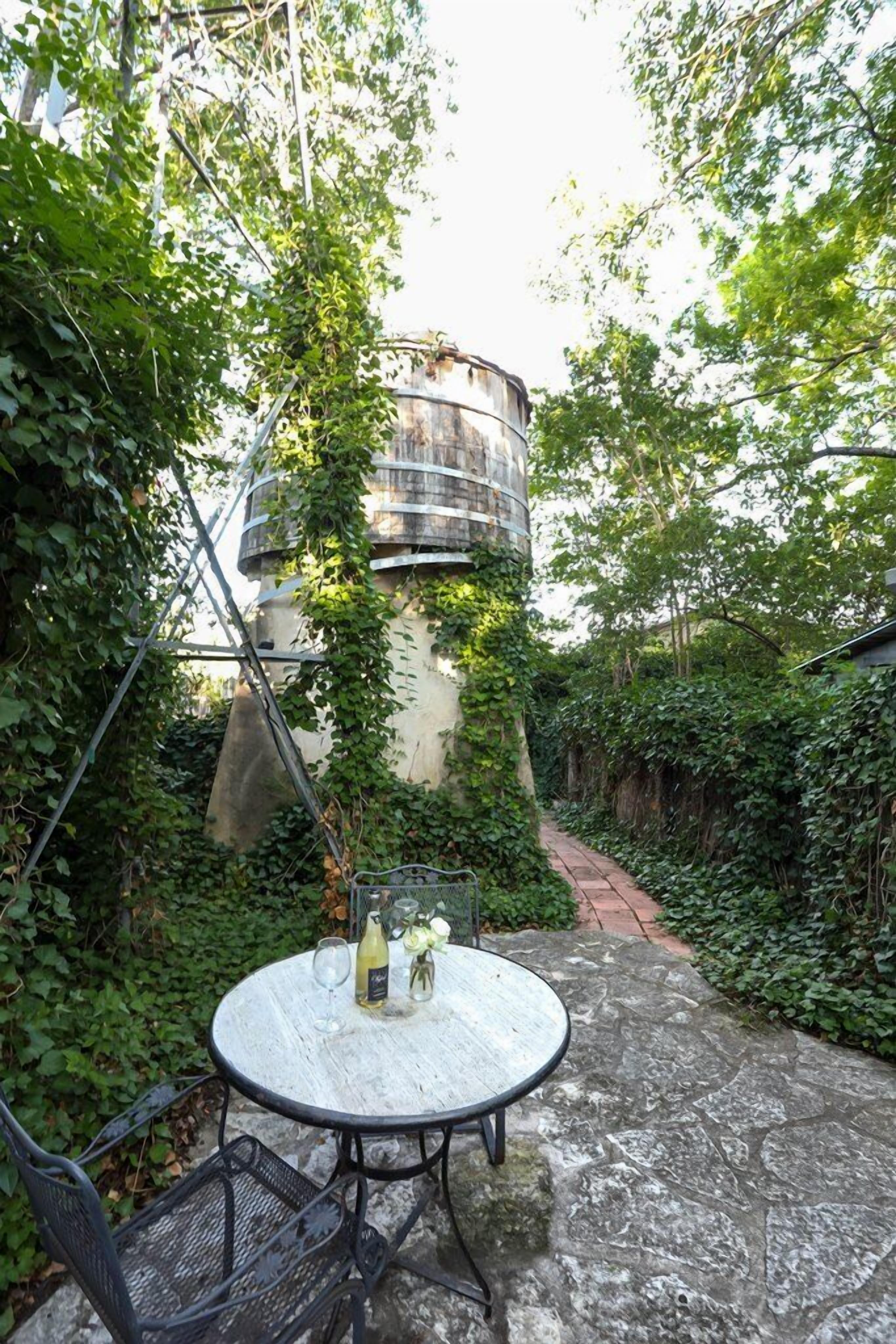 A round table with a metal frame and a white surface, surrounded by greenery and a stone pathway.