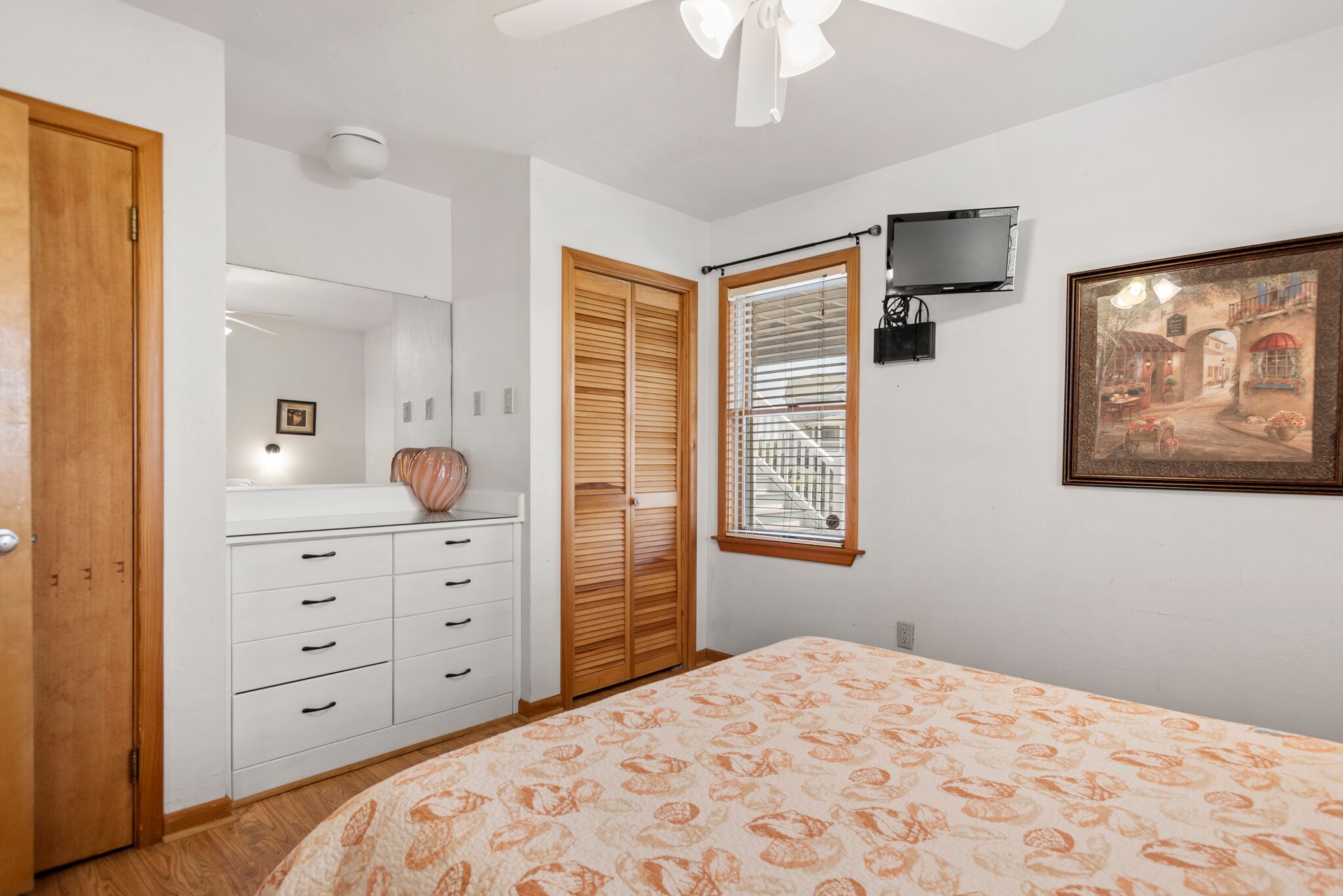 A bedroom with a bed, a wardrobe, a mirror, a window with blinds, a ceiling fan, a TV, and a framed picture on the wall.