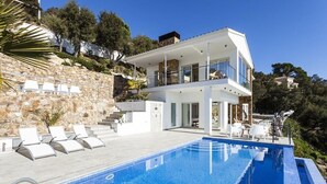 Una casa moderna de dos pisos con piscina, muros de piedra y un balcón con sillones de descanso.