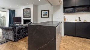 A modern living room with a dark sofa, a flat-screen TV, and a fireplace. The kitchen features dark cabinets and a marble countertop.