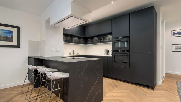 A modern kitchen with dark cabinetry, a central island with bar stools, and a herringbone wood floor.