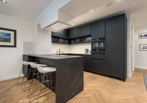A modern kitchen with dark cabinetry, a central island with bar stools, and a herringbone wood floor.