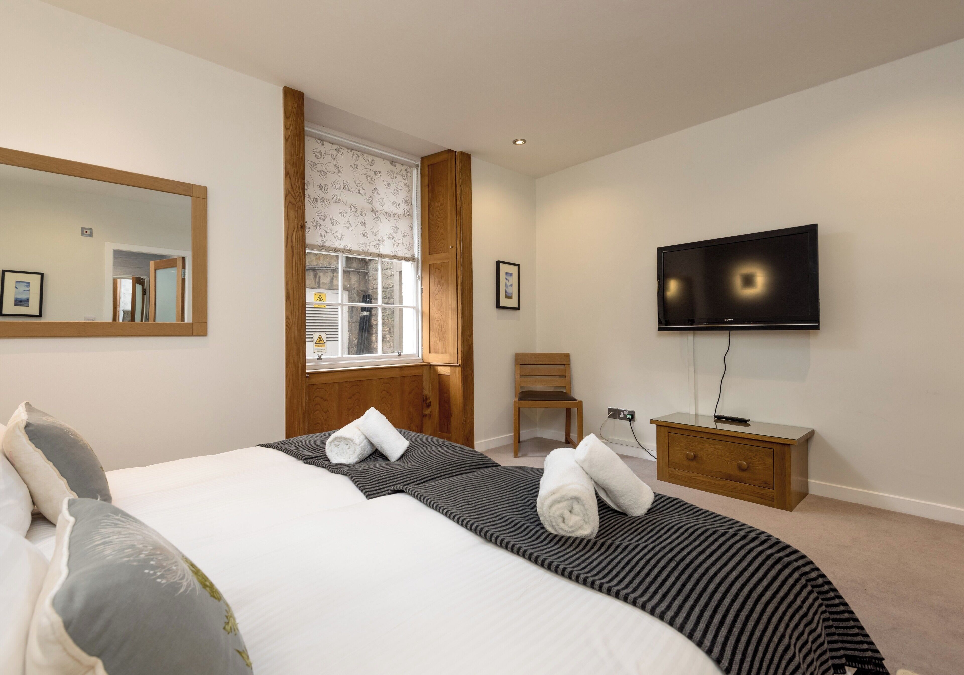 Habitación de hotel con cama, televisión, mesita de noche de madera, silla y ventana con cortinas.