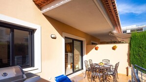 Une terrasse avec un barbecue, une table à manger avec des chaises, un fauteuil de jardin bleu et une haie verte.