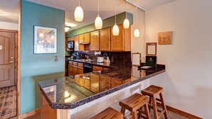 A kitchen with wooden cabinets, a granite countertop, hanging pendant lights, and a clock on the wall.