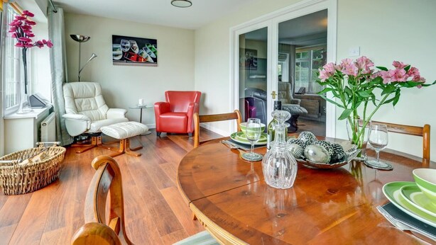 A dining area with a wooden table, chairs, a red chair, a white chair, a glass vase with flowers, and a painting on the wall. - 1 Bed in Northam, Bideford (oc-cedars) (Northam)