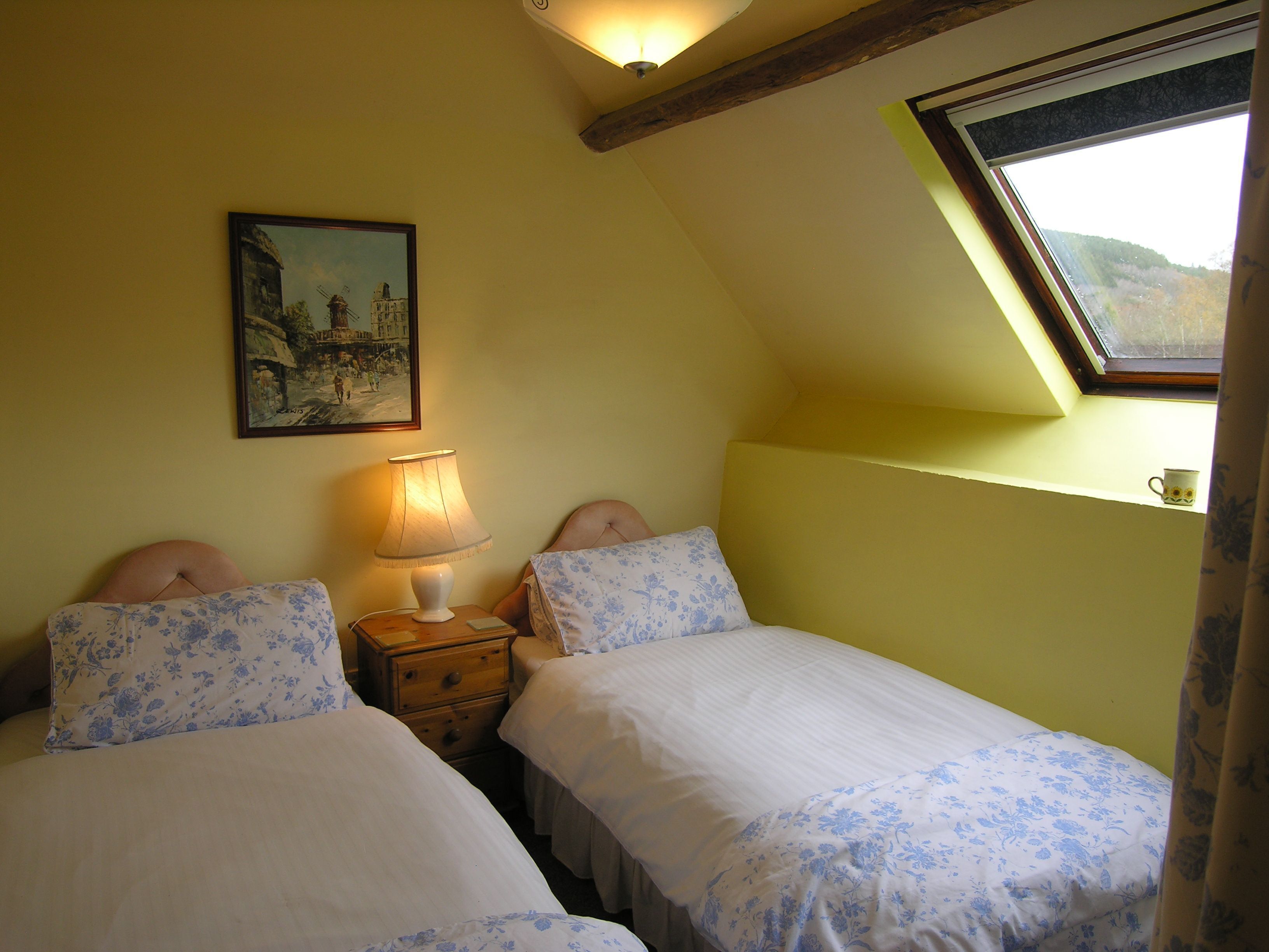 A bedroom with two beds, a nightstand, a lamp, a painting, and a skylight.