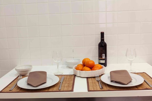 A dining table with two plates, two bowls, two wine glasses, a bottle of wine, and a bowl of oranges.