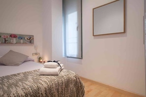 A bedroom with a bed, a window, a wall-mounted mirror, and a bedside table with a lamp.