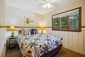 A bedroom with a bed, two bedside lamps, a ceiling fan, a framed picture, and a window with a view of trees. - 4-Bedroom Townhouse with Sauna - Flurry (Killington)