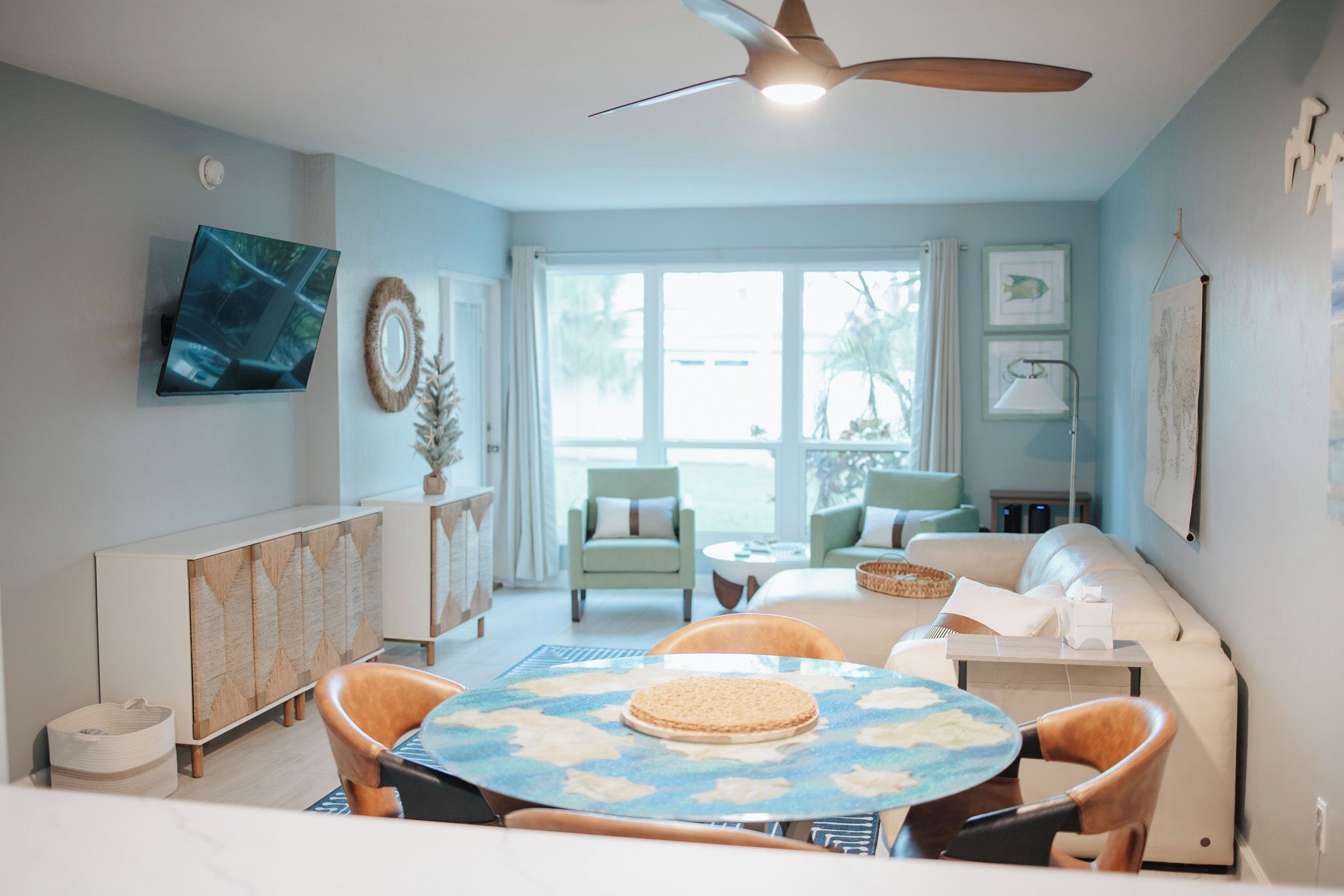 A living room with a round table, two chairs, a sofa, a ceiling fan, and a TV mounted on the wall.
