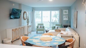 A living room with a round table, two chairs, a sofa, a ceiling fan, and a TV mounted on the wall. - Sunset Condo #156 on Anna Maria Island (Bradenton Beach)