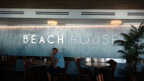 A restaurant interior with a wall displaying "BEACH HOUSE" in large letters, wooden tables, chairs, and a potted plant. - Sunset Condo #156 on Anna Maria Island (Bradenton Beach)