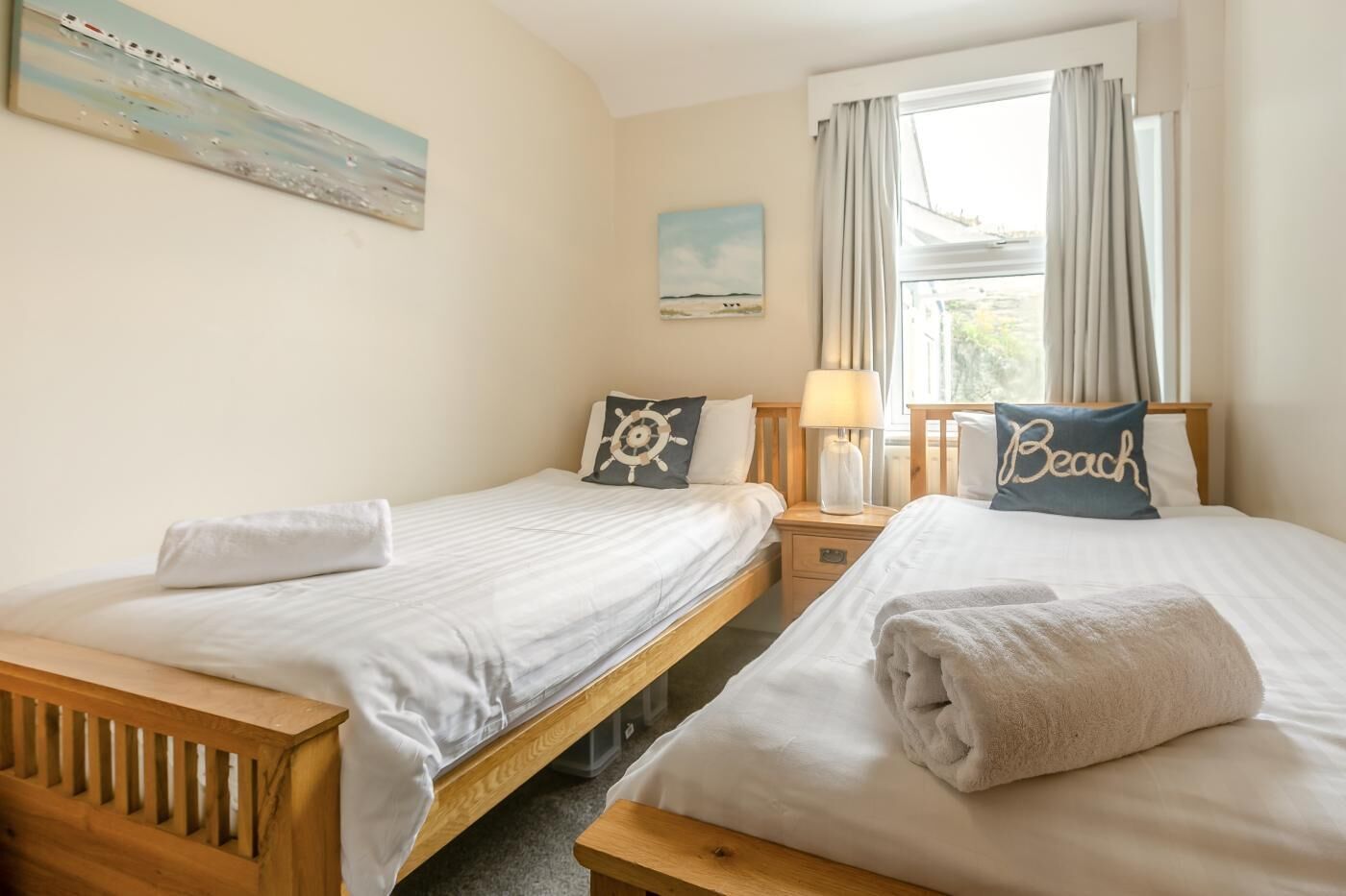 A hotel room with two beds, a nightstand with a lamp, a window with curtains, and artwork on the walls.