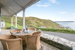 En overdækket terrasse med fletningsmøbler, et bord dækket med en croissant, kaffekopper og en tekedel.