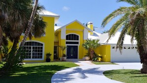 Une maison jaune avec une porte bleue, entourée de palmiers et d’une pelouse bien entretenue.