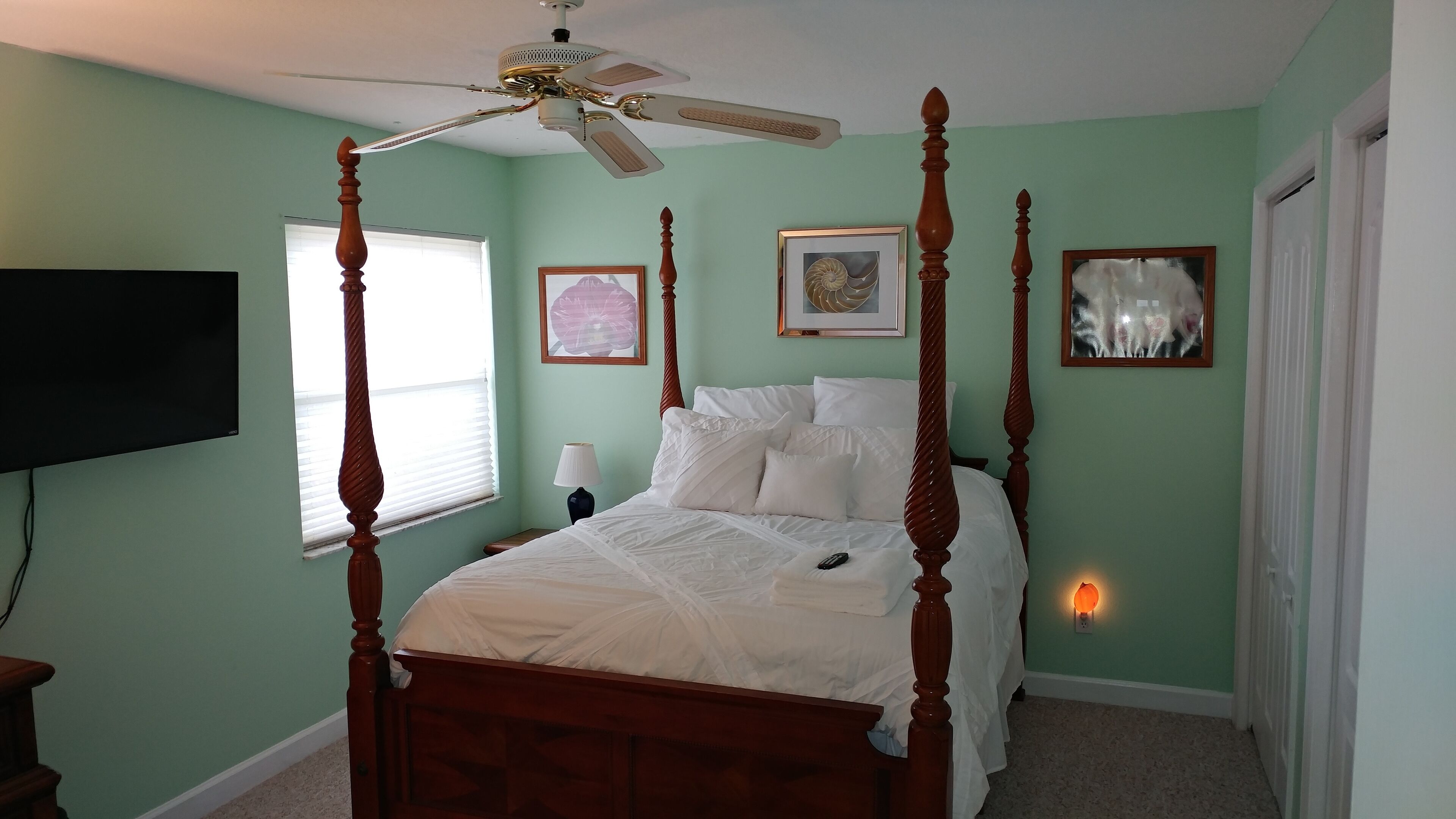 A four-poster bed with white linens, a ceiling fan, and framed artwork on the walls.