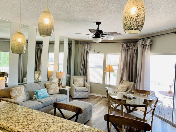 A spacious living area with a sofa, ottoman, and dining table with chairs. There are hanging lights and a ceiling fan.