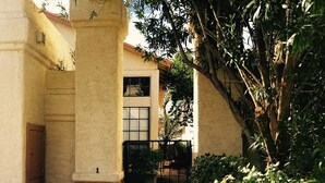 A building with a beige facade, a wooden door, and a black gate. There is a tree with green leaves to the right. - Tucson Omni Oasis – 4BR-3BA Desert Retreat with Private Hot Tub & Fire Pit (Tucson)