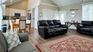 Amplia sala de estar con un sofá de cuero negro, un sofá gris y un tapete rojo estampado. Cuenta con un ventilador de techo, una mesa de comedor con sillas y una zona de cocina con armarios blancos y un microondas.