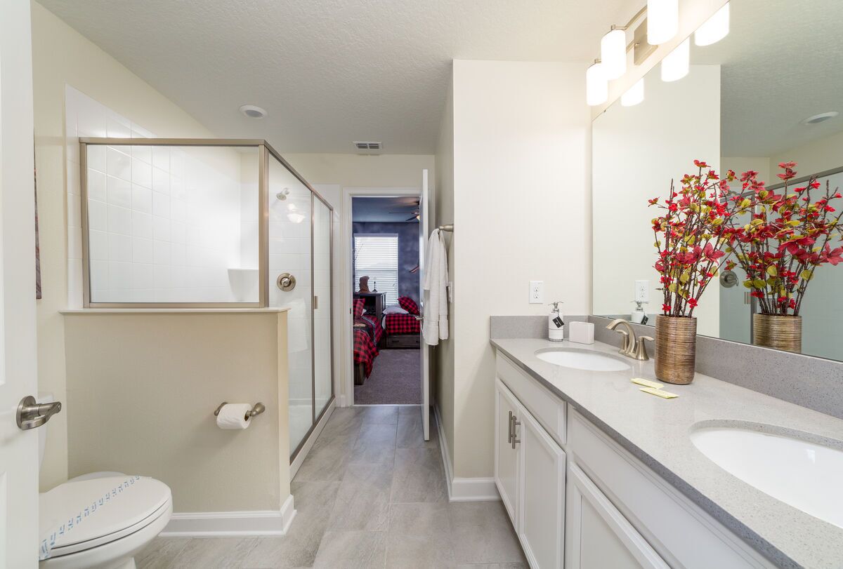 Een moderne badkamer met een dubbele wastafel, toilet en een grote spiegel. Er is een gang die leidt naar een slaapkamer met een rood en zwart geruit beddegoed.