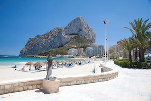 A beach with a rocky mountain in the background, a walkway with umbrellas, and buildings nearby. - Villa Alaska - PlusHolidays (Calpe)