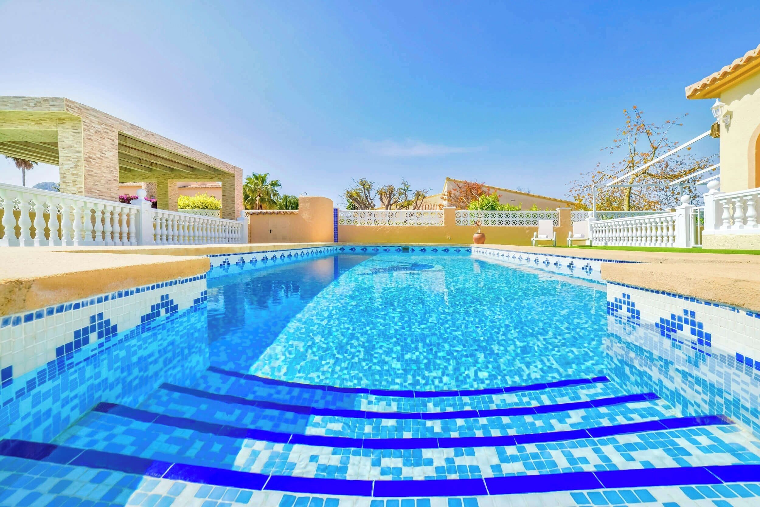 A swimming pool with clear blue water, surrounded by a white fence and a covered area with plants.