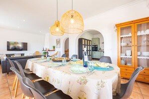 A dining area with a round table set for four, surrounded by black chairs. There are two hanging wicker pendant lights above the table. A wooden cabinet with glass doors is to the right, containing plates and glasses.