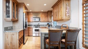 A kitchen with wooden cabinets, a stainless steel refrigerator, a microwave, and a sink. There is a bar area with two chairs and a wine bottle. - Townhome Near Main St | Skiing | Hot Tub (Park City)