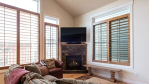 A cozy living room with a fireplace, large windows with blinds, a leather sofa, and a stone accent wall. - Townhome Near Main St | Skiing | Hot Tub (Park City)