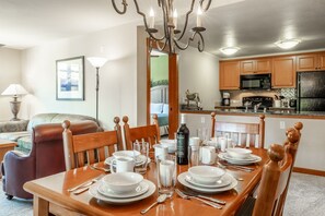 Un comedor con una mesa de madera preparada para cuatro personas, una araña y una cocina al fondo.