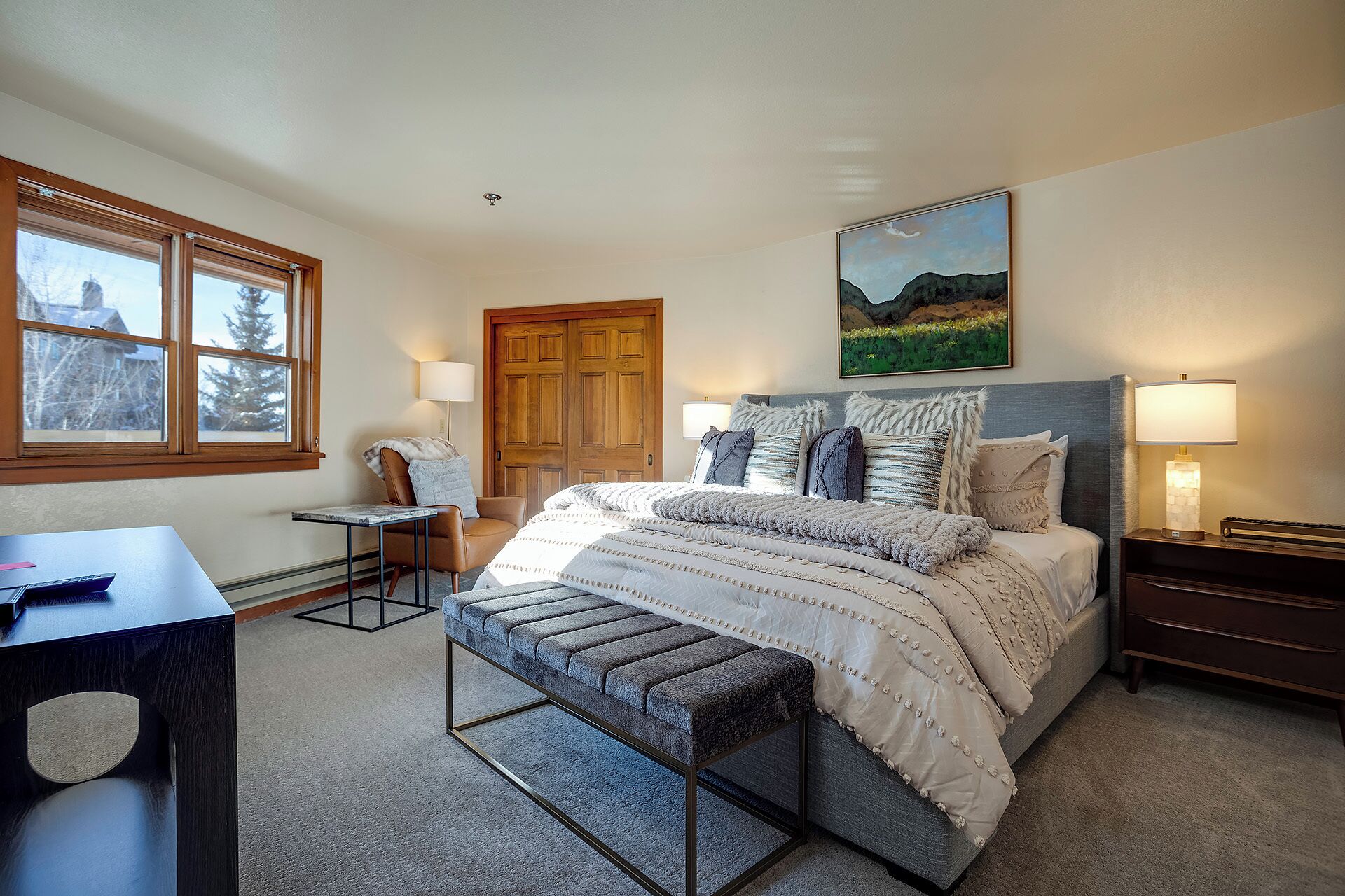 A bedroom with a bed, a desk, a chair, a bench, a door, and a painting on the wall.