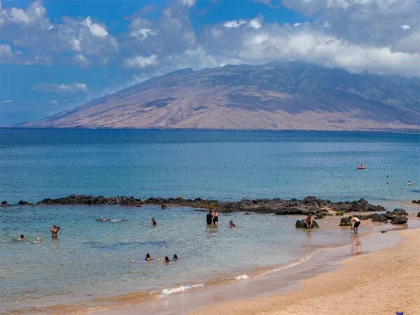 A beach with clear water, rocky areas, and a mountain in the background.
