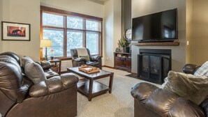 A living room with leather sofas, a coffee table, a fireplace, a television, and a large window with blinds. - Solstice 21 | Easy Walk to Eagle Lodge (mammoth lakes)