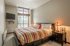 Ein Schlafzimmer mit einem Bett, Nachttischlampen, einem an der Wand montierten Fernseher und einem Fenster mit Jalousien.