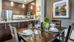 A modern kitchen with dark wood cabinets, stainless steel appliances, and a dining area set for six with a large table, chairs, and a vase of flowers. - Solstice 21 | Easy Walk to Eagle Lodge (mammoth lakes)