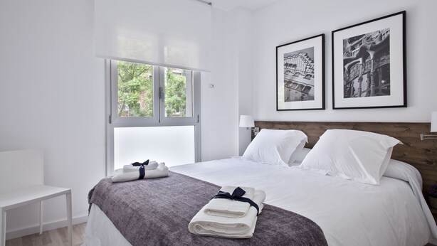 A modern hotel room with a large bed, a bedside table, a window with a view of greenery, and two framed pictures on the wall. - Blanca 1 - Habitat Apartments (Barcelona)