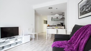 A modern living room with a flat-screen TV, a black sofa with a purple throw, and a dining area with a white table and chairs. - Blanca 1 - Habitat Apartments (Barcelona)