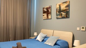 A hotel room with a bed, two pillows, a blue bedspread, a wooden headboard, two wall-mounted paintings, a window with curtains, and a bedside table with a lamp.