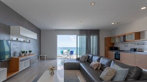 A modern living room with a large sofa, a coffee table, and a TV. There is a kitchen area with cabinets and a dining area with a table and chairs.