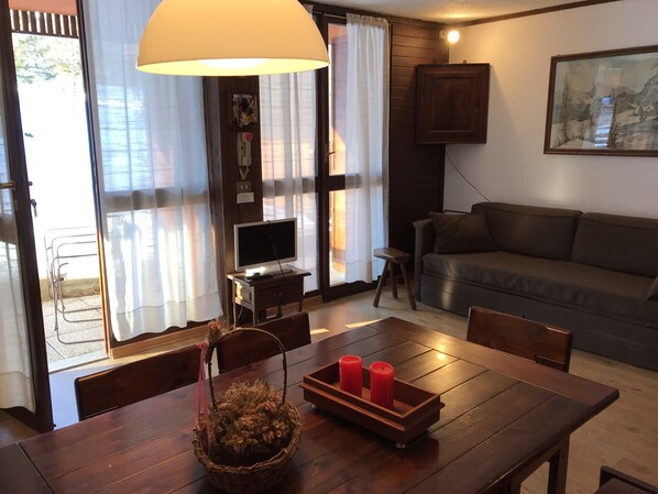 A living room with a sofa, a TV on a stand, a dining table with chairs, a lamp, and a painting on the wall.