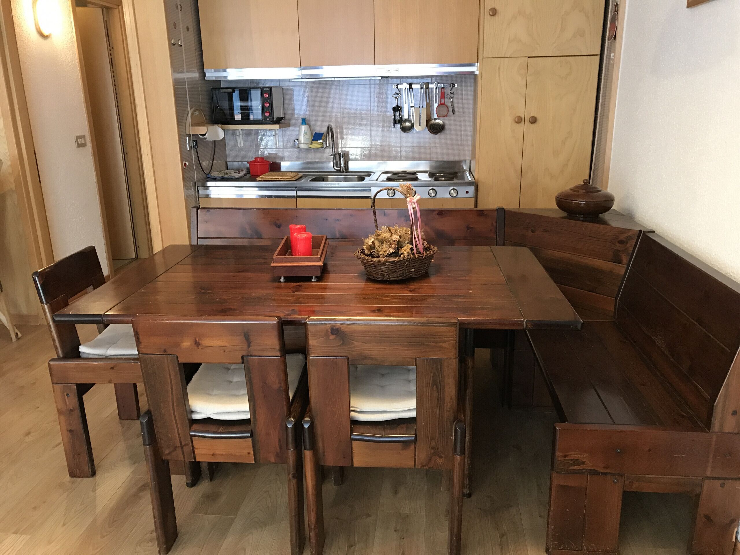 Une table à manger en bois avec des bancs, un coin cuisine équipé d’un four à micro-ondes, d’une cuisinière et d’ustensiles de cuisine suspendus au-dessus de l’évier.