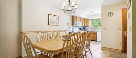 Un comedor con mesa y sillas de madera, una araña y una cocina al fondo.