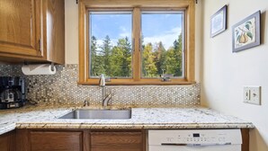 Una cocina con armarios de madera, un lavamanos de acero inoxidable, un lavavajillas Bosch y un ventanal con vista a árboles.