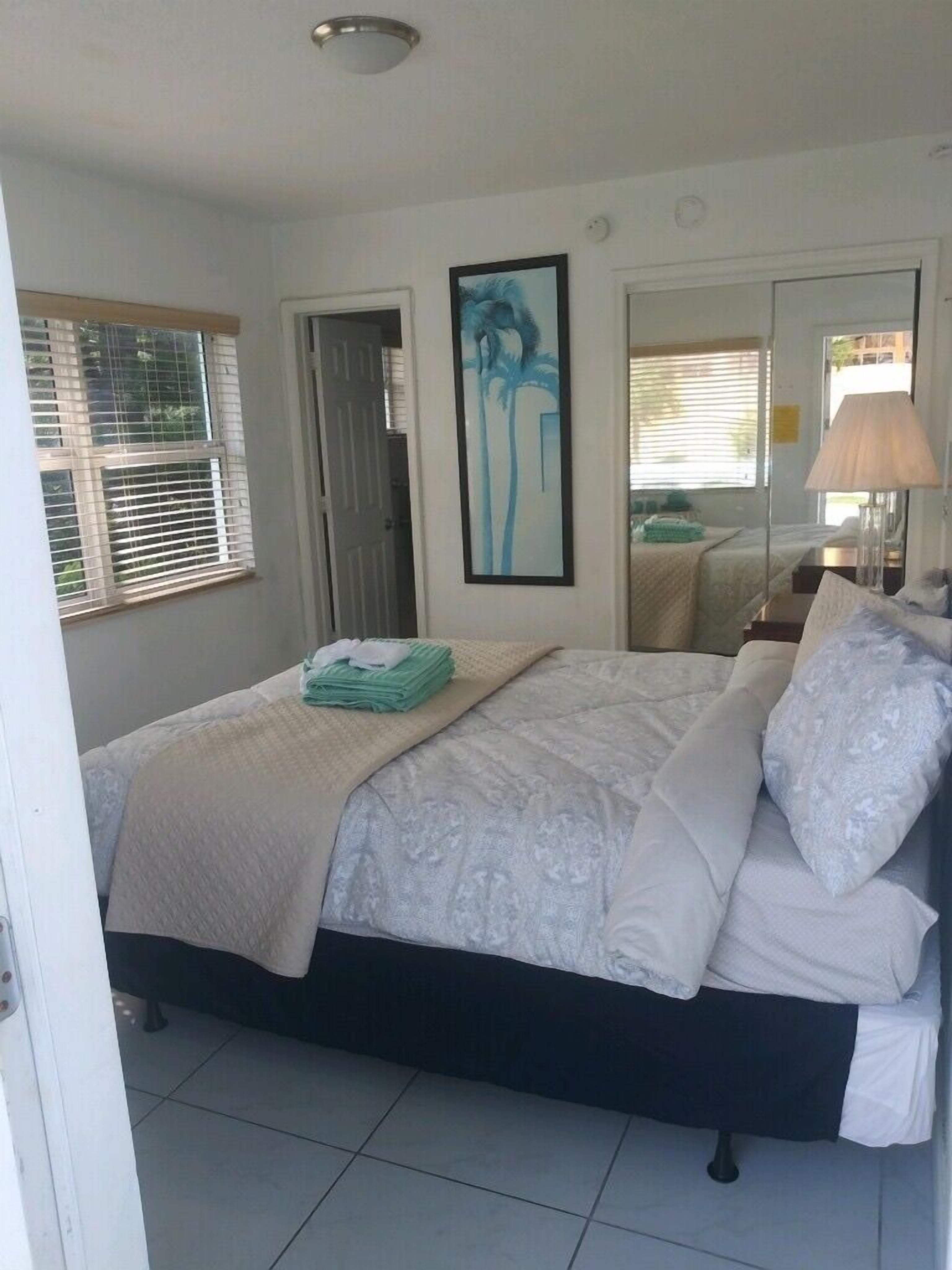 A neatly made bed with a white and blue patterned bedspread, a folded green towel, and a white pillow. There is a framed picture on the wall above the bed, a window with blinds, and a mirror reflecting part of the room.