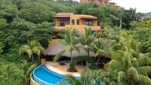 A resort with a pool surrounded by palm trees and a thatched-roof structure. - Villa Encantada at Pacific Vacation | StayPV Oceanfront Luxury with Staff (Zihuatanejo)