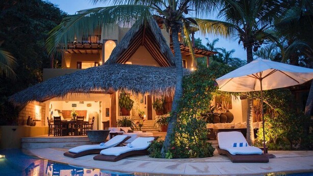 A resort with a pool, lounge chairs, and a thatched-roof structure. - Villa Encantada at Pacific Vacation | StayPV Oceanfront Luxury with Staff (Zihuatanejo)