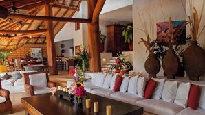 A spacious living area with a wooden ceiling, white sofas with red cushions, a dark wooden coffee table, and decorative vases. - Villa Encantada at Pacific Vacation | StayPV Oceanfront Luxury with Staff (Zihuatanejo)